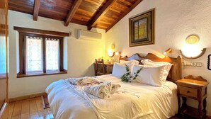Una camera da letto con soffitto in legno, un letto con lenzuola bianche, un comodino, una lampada a parete e una finestra con tende.