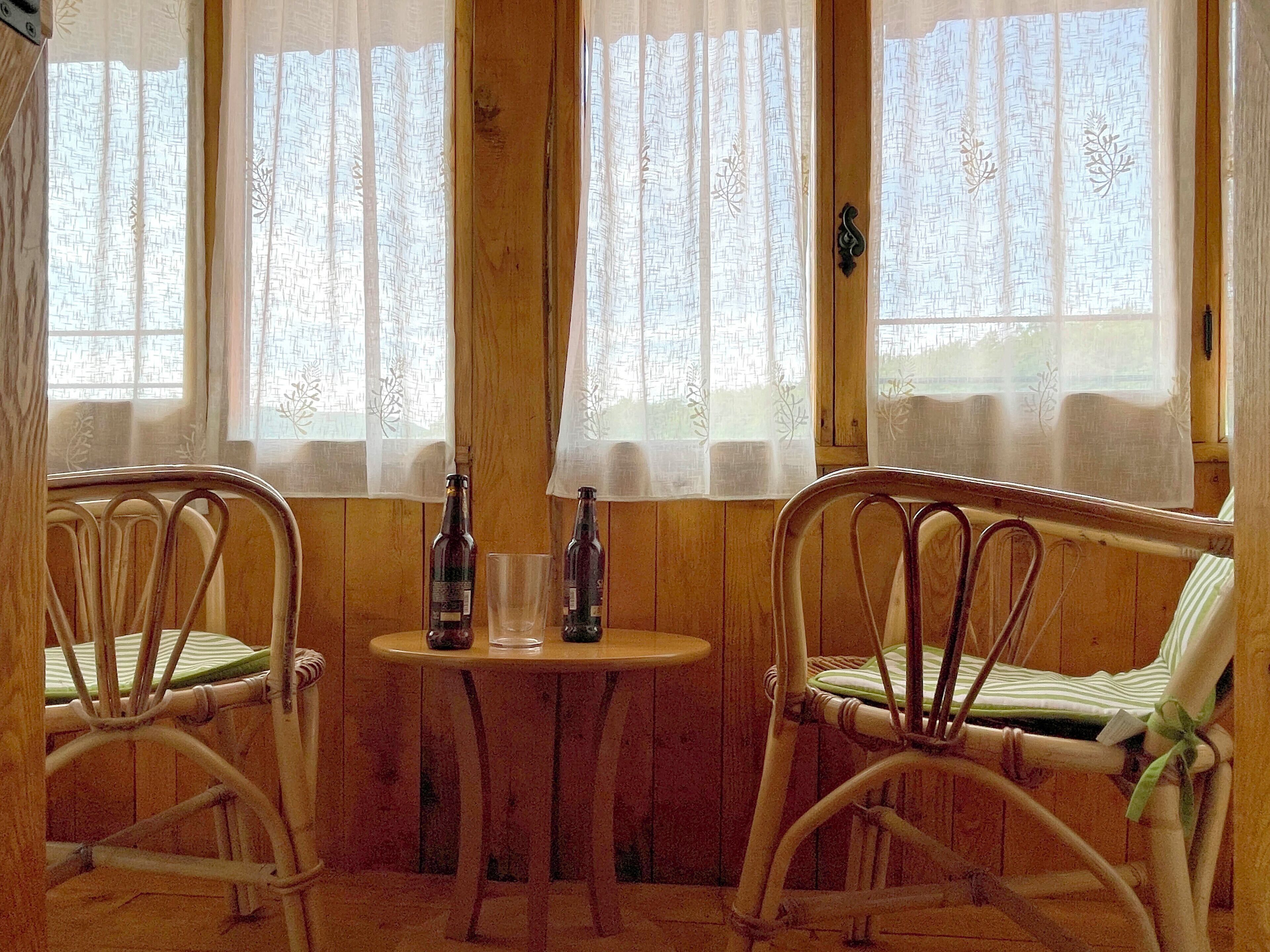 A cozy room with wooden walls and two wicker chairs facing a small table with two bottles and a glass.