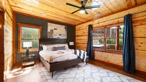 Camera da letto in una baita di legno con un letto, una ventola a soffitto, una finestra con tende e vista sull'esterno.