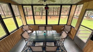Un patio techado con una mesa y sillas de vidrio, un ventilador de techo y vista a un jardín cubierto de césped.