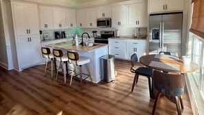 A modern kitchen with white cabinets, a stainless steel refrigerator, and a dining area with wooden chairs. - The Bay Stay has boat pier ~ 4 mi. to Beach. Cart into town- Close to Boat ramp  (Bay St. Louis)