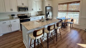 A spacious kitchen with white cabinets, a central island with bar stools, a stainless steel refrigerator, and a large window providing natural light. - The Bay Stay has boat pier ~ 4 mi. to Beach. Cart into town- Close to Boat ramp  (Bay St. Louis)