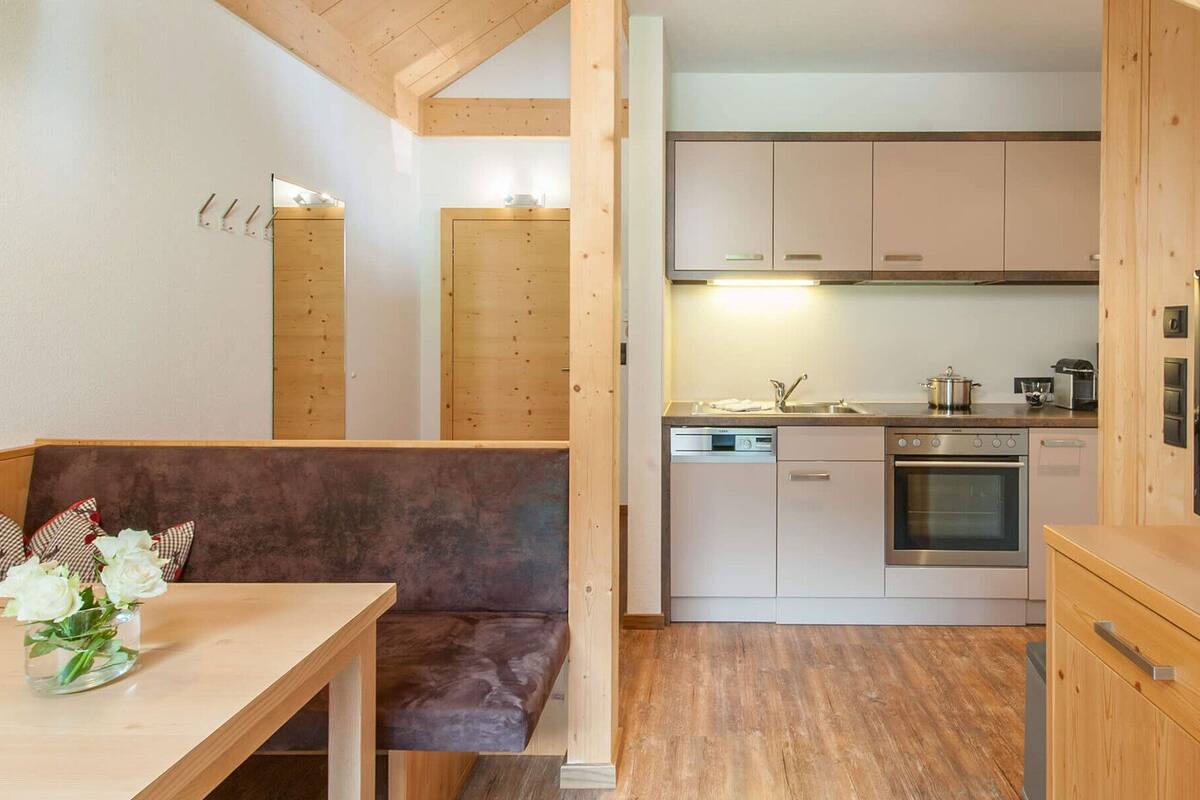 Une cuisine moderne avec des armoires en bois, un four encastré et un lave-vaisselle. On y trouve un coin repas avec un banc et une table, et un téléviseur sur le côté droit.