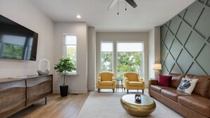 A modern living room with a wooden floor, a large TV on the wall, two yellow armchairs, a brown leather sofa, and a herringbone-patterned rug.