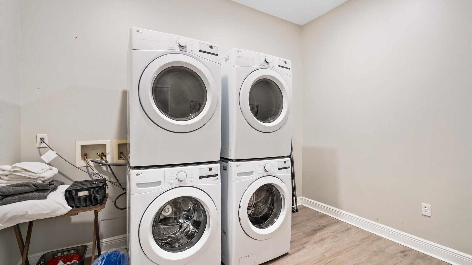 A laundry room with two stacked front-loading LG washing machines, a drying rack with folded clothes, and a utility area with a water heater and electrical outlet.