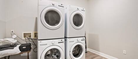 A laundry room with two stacked front-loading LG washing machines, a drying rack with folded clothes, and a utility area with a water heater and electrical outlet.