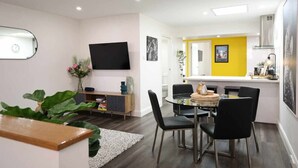 Una sala moderna con un televisor de pantalla plana, una zona de comedor con una mesa redonda y sillas negras, y una cocina con una pared de acento amarilla.