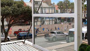 Vista desde una ventana con piso de metal corrugado, que da a una calle con coches aparcados y un mural en un edificio.
