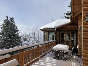 Una cabaña de madera con un porche cubierto y una mesa con sillas, rodeada de árboles cubiertos de nieve.
