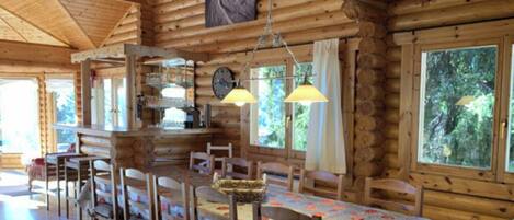 A wooden cabin with a dining area featuring a table and chairs, a kitchen area with a clock, and large windows offering views of the outdoors.