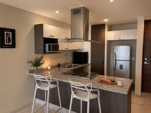Et moderne køkken med en central ø, rustfrie hvidevarer og hvide barstole.