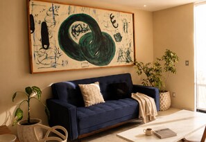 Une pièce avec un canapé bleu foncé, un grand tableau abstrait, deux plantes en pot et une table sur laquelle se trouvent un livre et une tasse.