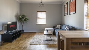 Ein Wohnzimmer mit Fernseher, der 'NETFLIX' anzeigt, einer Couch, einem Couchtisch, einem Topf mit Pflanze und gerahmten Kunstwerken an der Wand.