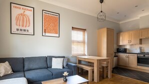 Ein modernes Wohnzimmer mit grauem Sofa, einem Holztisch und einem Essbereich mit Holztisch und Bänken. Es gibt eine Küche mit Holzschränken und einem schwarzen Herd. Zwei gerahmte Poster hängen an der Wand.