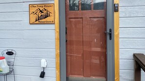 A wooden cabin with a brown door, a welcome mat, a sign reading "The Tiny House Walnut Grove B," and a small outdoor seating area. - Tiny House on Walnut Grove (Ashland)