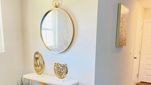 Un pasillo con una consola blanca, adornos dorados, un espejo redondo y una obra enmarcadas en la pared.