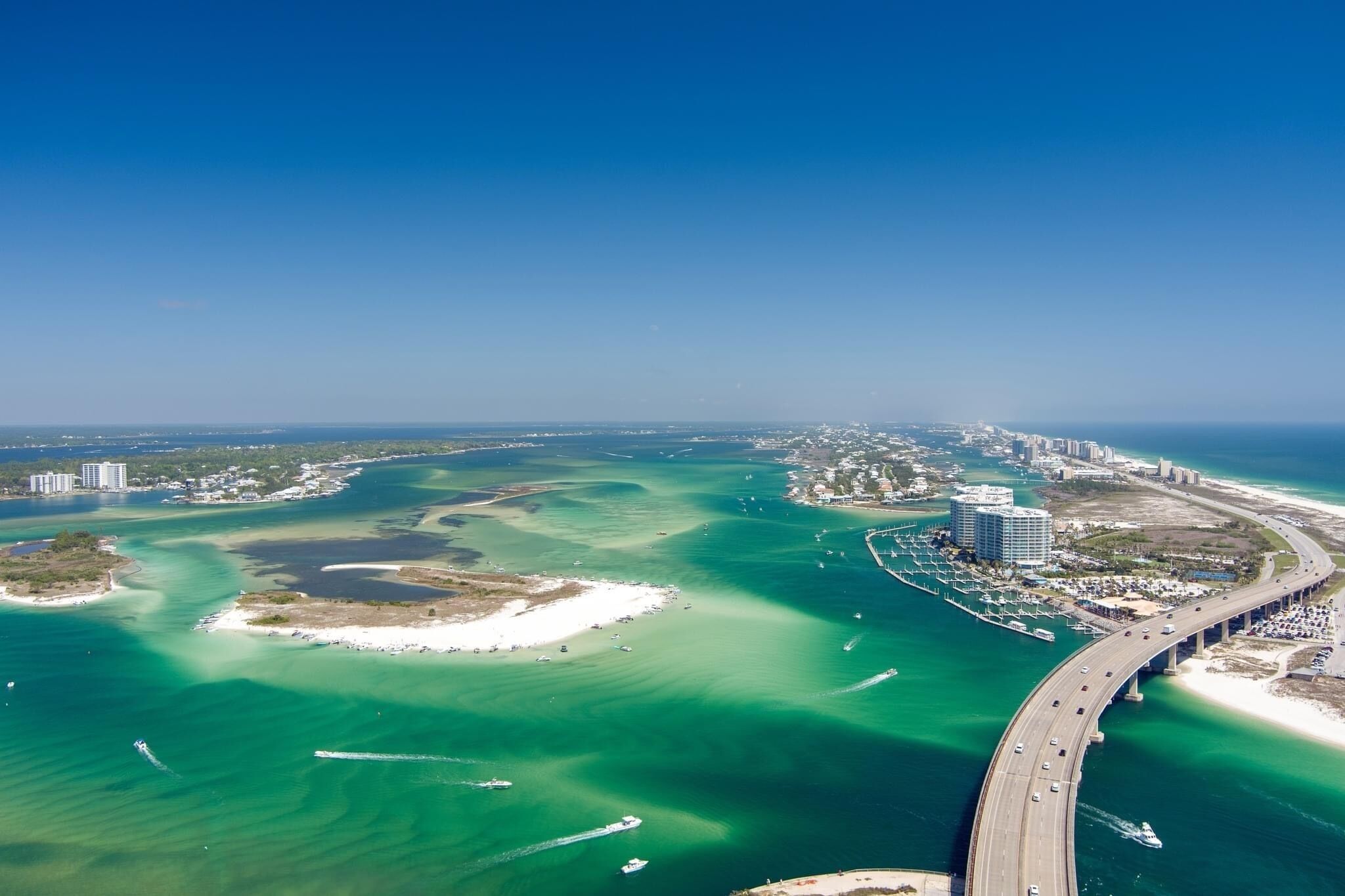 An aerial view of a coastal city with a prominent bridge, clear blue waters, and a mix of urban and natural landscapes.