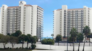 Two high-rise buildings with balconies, a clear sky, and a parking area with palm trees. - Amazing Corner Views, Huge Balcony on the Beach! Newly Remodeled Inside and Out! (Orange Beach)