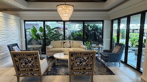 A spacious living room with a beige sofa, two wooden chairs, a coffee table, and a chandelier. Large windows offer a view of greenery outside. - Marco Island Vacation Rental, Beachfront Condo with Unobstructed Gulf Views,Pool (Marco Island)