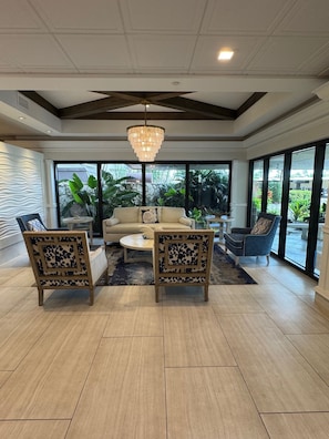 A spacious living room with a beige sofa, two wooden chairs, a coffee table, and a chandelier. Large windows offer a view of greenery outside. - Unobstructed Gulf Views From All Rooms & Balcony + Direct Beach Access ! (Marco Island)