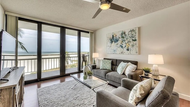 A spacious living room with a large sliding glass door leading to a balcony, a ceiling fan, a gray sofa set, and a painting on the wall. - Marco Island Vacation Rental, Beachfront Condo with Unobstructed Gulf Views,Pool (Marco Island)