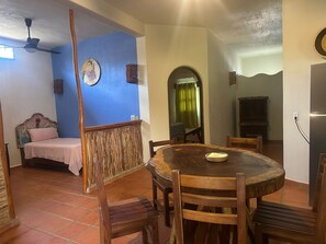 Un coin repas avec une table ronde en bois, des chaises, un lit avec une tête de lit, un ventilateur de plafond et un mur orné d’une œuvre d’art circulaire.