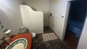 Et badeværelse med toilet, håndvask, brusebad, og en seng i det tilstødende rum.