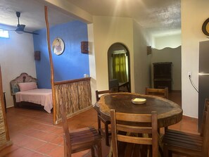 A dining area with a round wooden table, chairs, a bed with a headboard, a ceiling fan, and a wall with a circular artwork. - Habitación Casa Flores (Planta baja) (San Francisco)