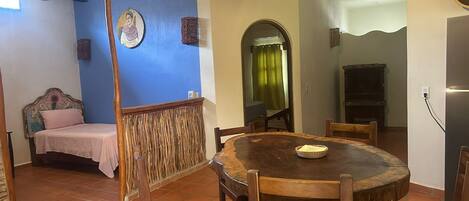 Un comedor con una mesa redonda de madera, sillas, una cama con cabecera, un ventilador de techo y una pared decorada con una obra de arte circular.