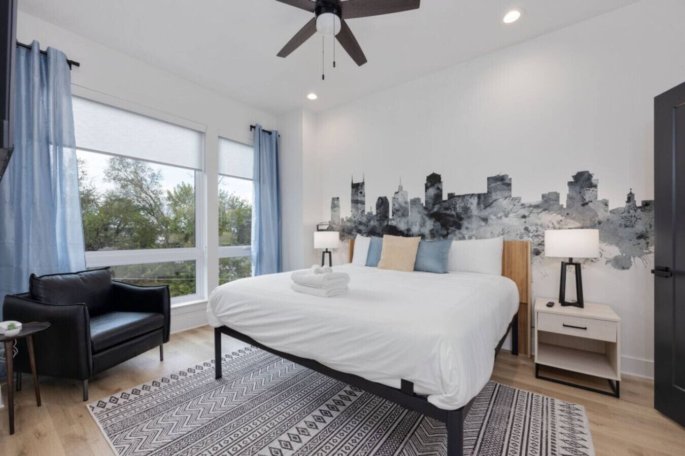 Quarto moderno com uma cama grande, um sofá de couro preto, uma obra de arte com paisagem urbana e vista para a cidade pela janela.