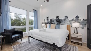 A modern bedroom with a large bed, a black leather sofa, a cityscape wall art, and a city view through the window. - `The Ryman` 4BR Haven with Scenic Rooftop (Nashville)