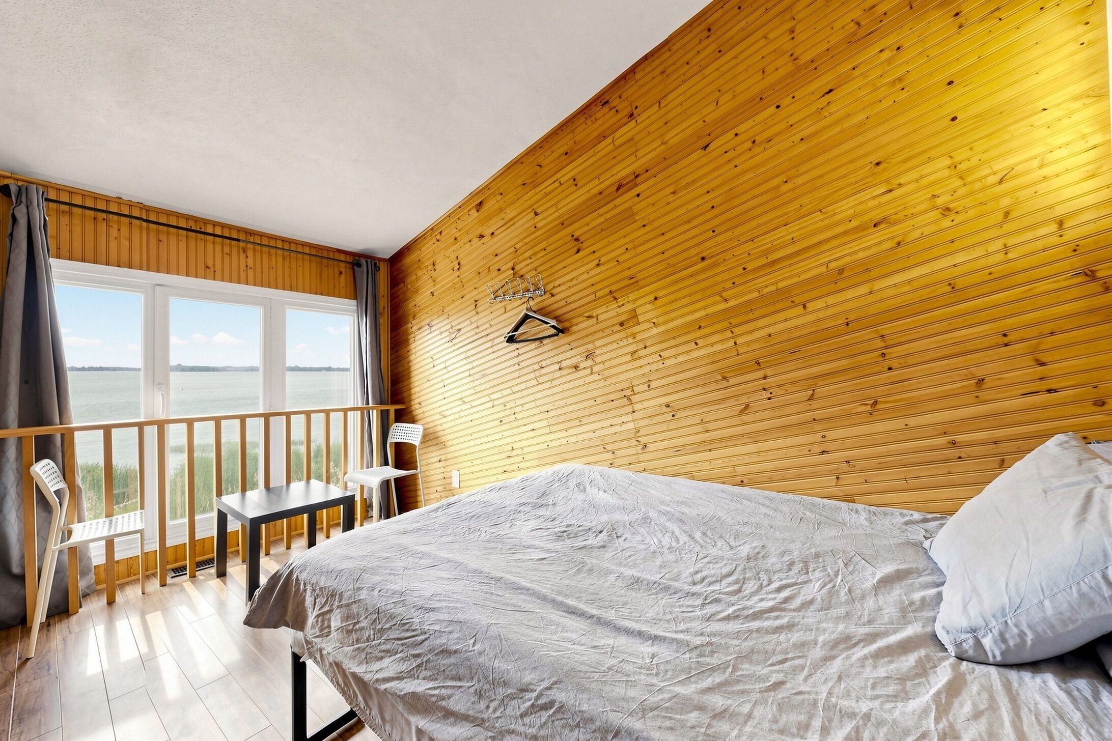 A bedroom with a bed, a small table, a chair, a balcony with a railing, and a view of the outdoors.