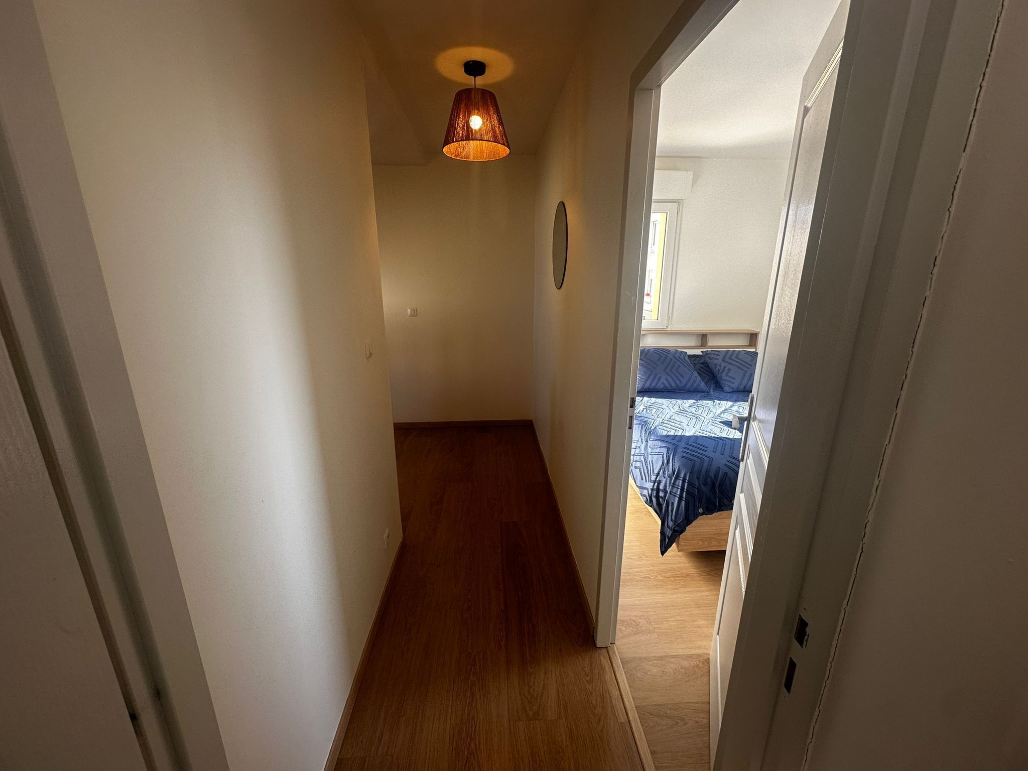 A narrow hallway with wooden flooring leading to a bedroom with a bed and a hanging light fixture.