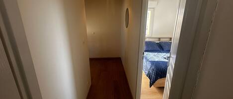 A narrow hallway with wooden flooring leading to a bedroom with a bed and a hanging light fixture.