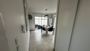 A hallway leading to a dining area with a table and chairs, a clock on the wall, and a window with a view of the outside.
