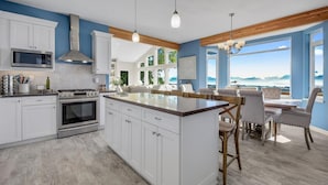 A modern kitchen with white cabinetry, a central island, and a dining area with a view. - Spacious home w full Kachemak Bay views (Homer)
