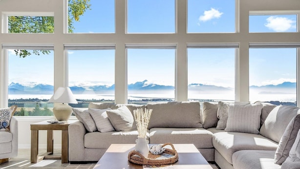 A modern living room with a large sectional sofa, a coffee table, and a view of mountains through large windows. - Spacious home w full Kachemak Bay views (Homer)