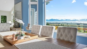 A modern living room with a large window offering a mountain view, a wooden dining table, and comfortable seating. - Spacious home w full Kachemak Bay views (Homer)