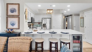 A modern kitchen with white cabinets, a central island with stools, a refrigerator, and a beverage cooler. - Emerald Escape! Newly remodeled spacious 3-bedroom condo with oversized balcony! (Destin)