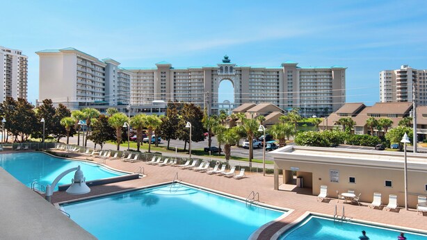 A hotel with multiple swimming pools, palm trees, and a large building with a distinctive archway in the background. - Emerald Escape! Newly remodeled spacious 3-bedroom condo with oversized balcony! (Destin)
