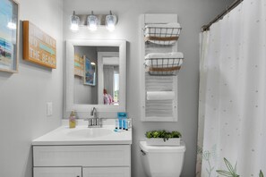 A bathroom with a white sink, mirror, and toilet. There are three light fixtures above the mirror, a towel rack with multiple towels, and a small plant on the toilet. - Emerald Escape! Newly remodeled spacious 3-bedroom condo with oversized balcony! (Destin)