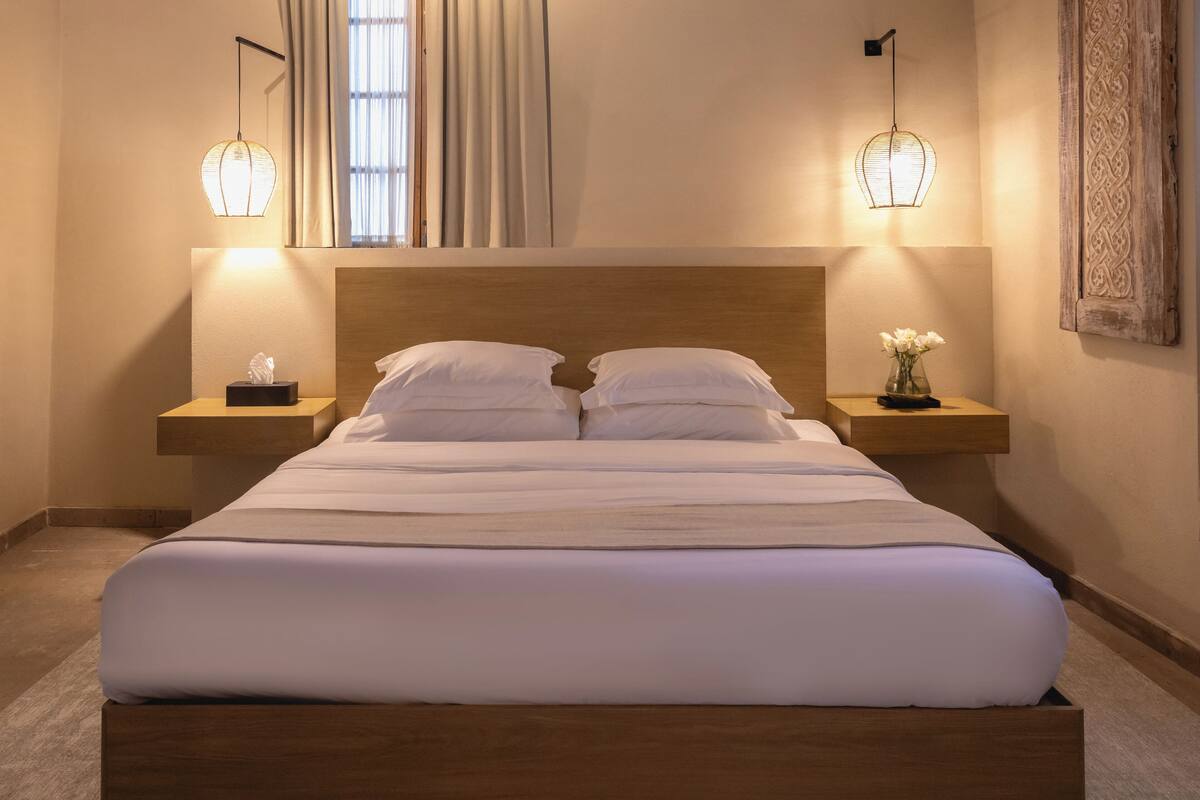 Una cama bien hecha con sábanas blancas, dos mesitas de noche con lámparas, un jarrón con flores y un espejo sobre la cama.