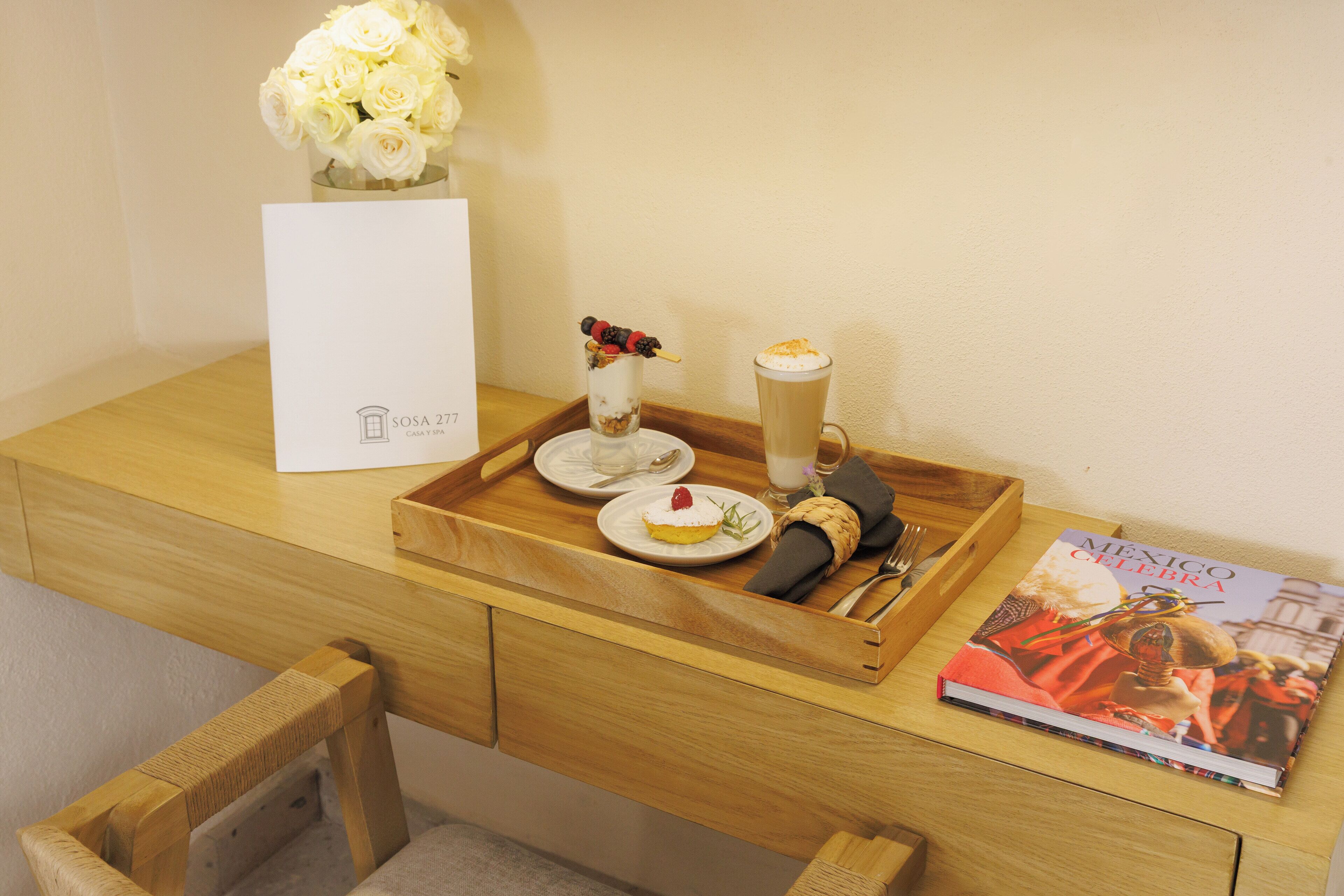 A wooden desk with a tray holding a dessert, a book titled 'Mexico Celebrates', and a vase with white flowers.
