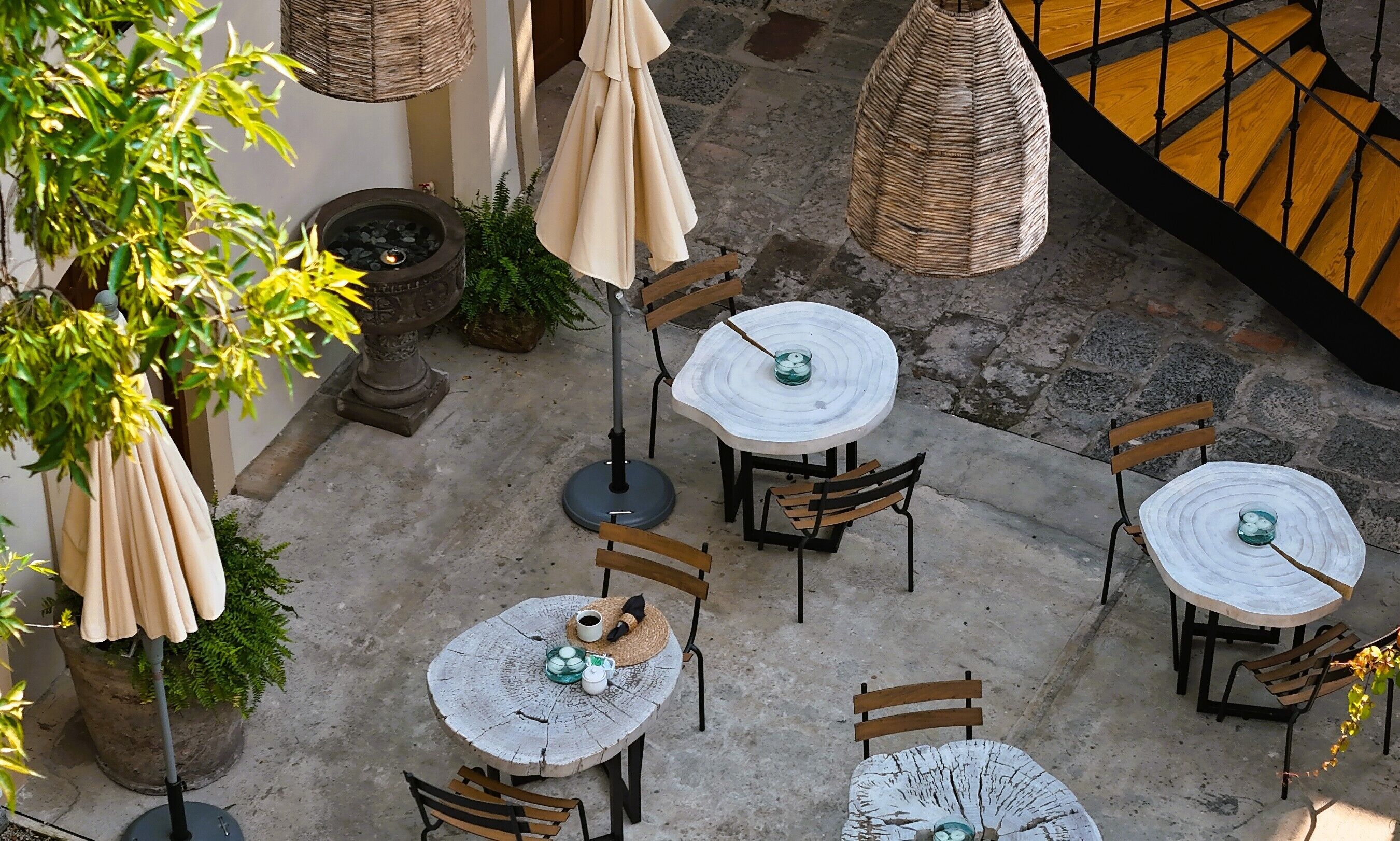 Ein Terrassenbereich im Freien mit runden Tischen und Stühlen, umgeben von Blumentöpfen und hängenden Korbuschirm