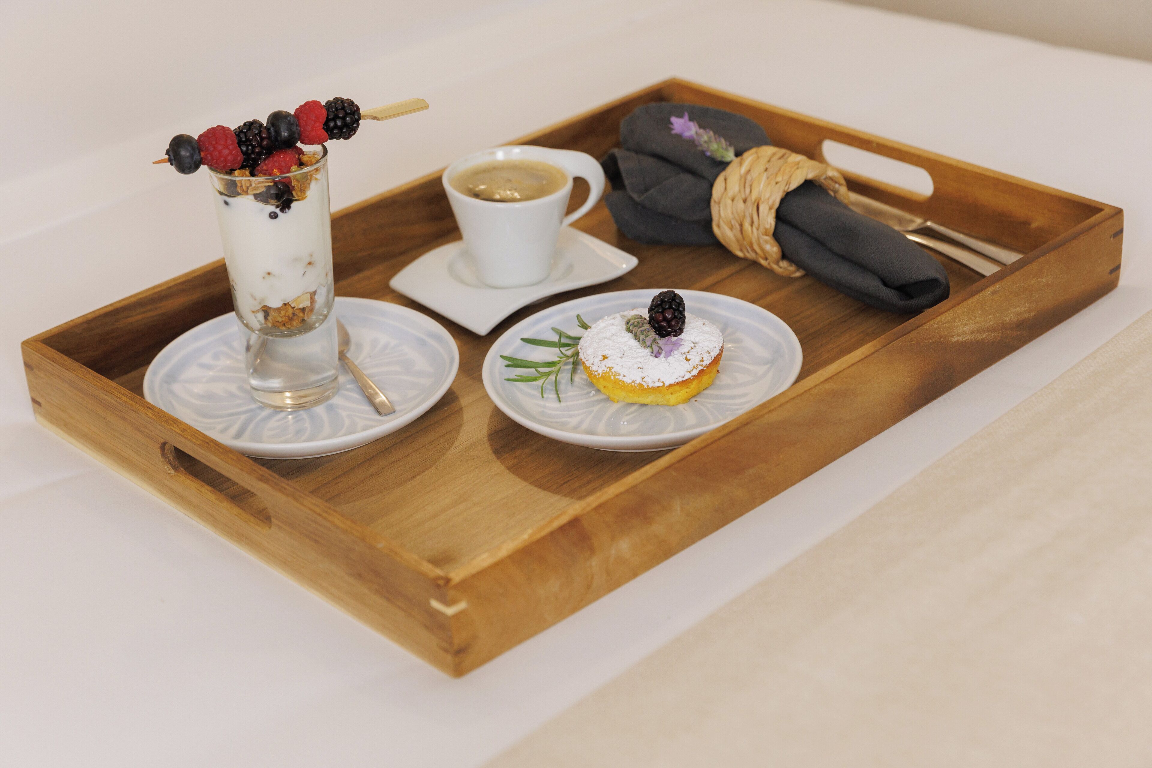 A wooden tray with a glass of yogurt parfait, a cup of coffee, a wrapped napkin, and a plate with a pastry.