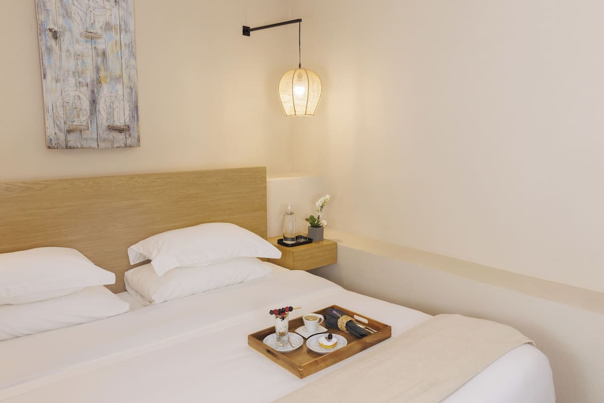 Una cama bien tendida con ropa blanca, un cabecero de madera, una mesita de noche con una lámpara y una bandeja con alimentos para el desayuno.