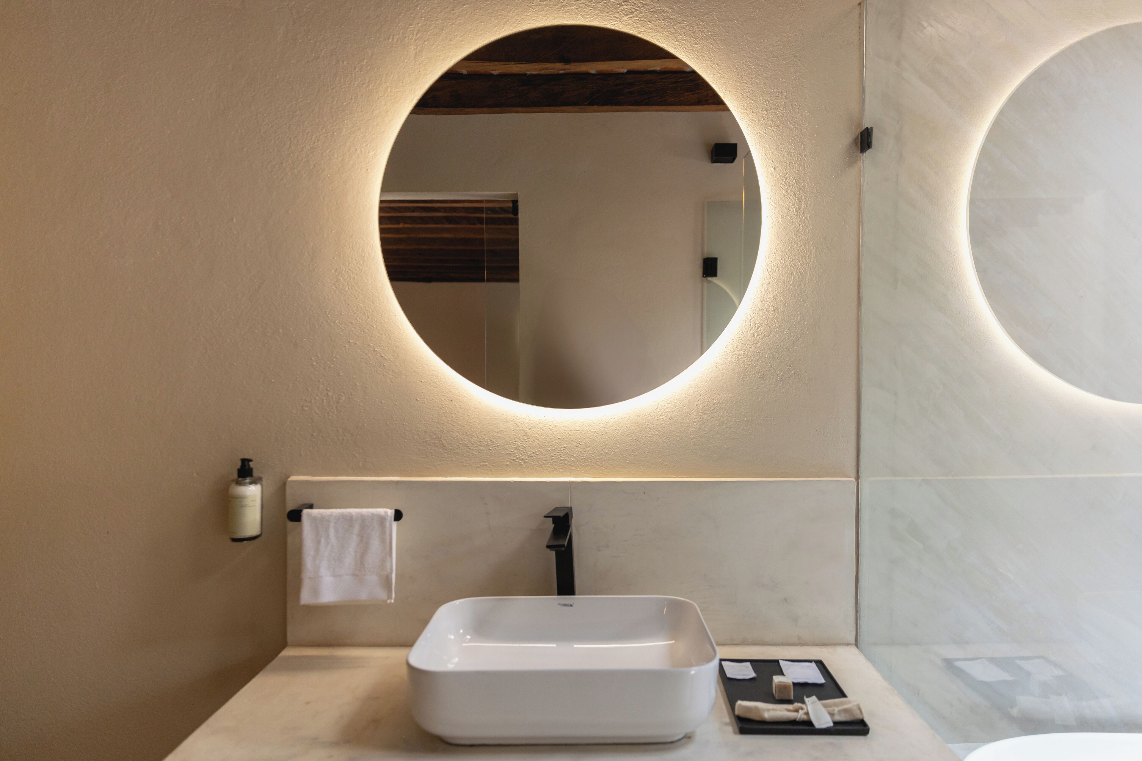 Un baño moderno con un lavamanos rectangular, un espejo redondo y una toalla colgada en la pared.