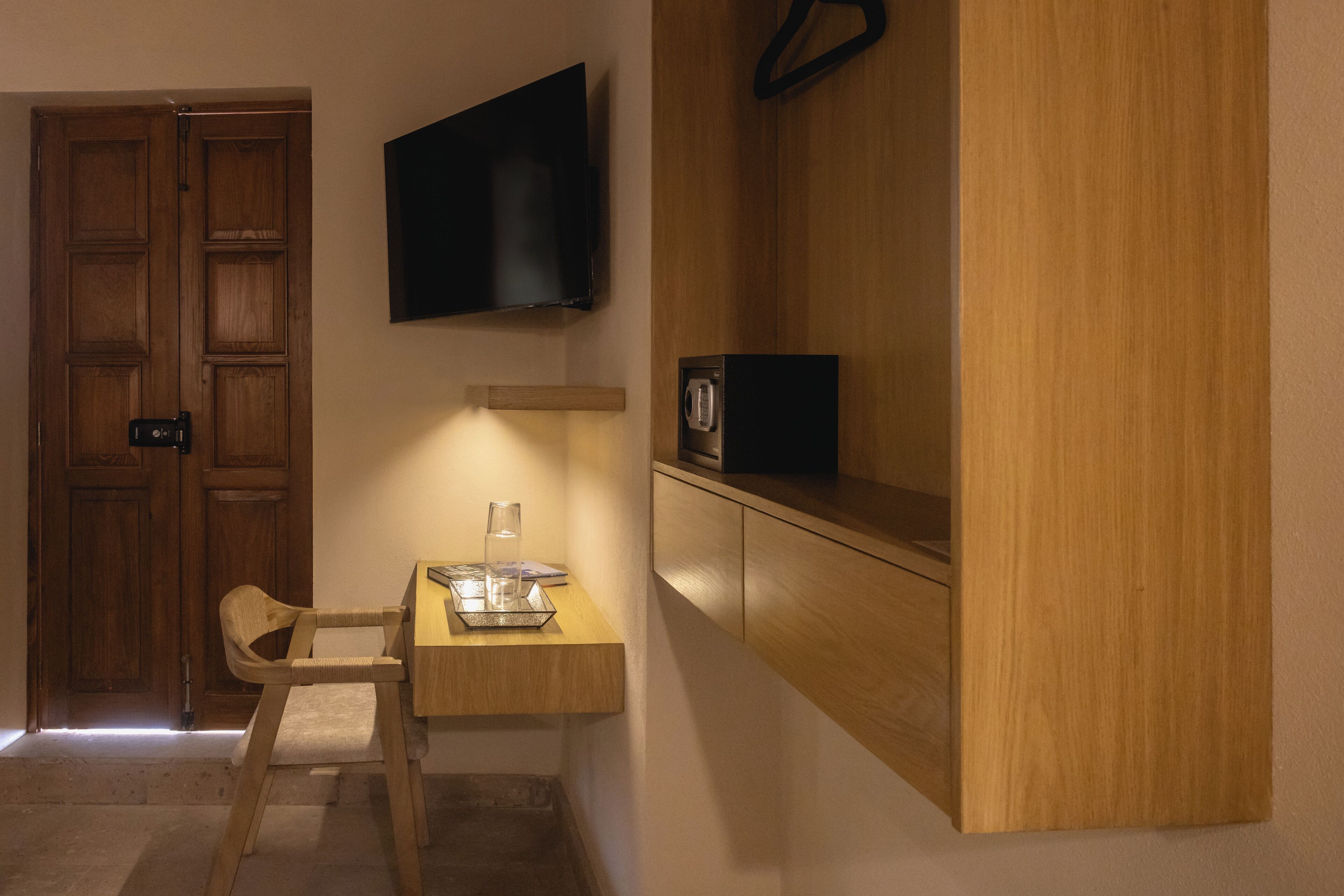 Una habitación con puerta de madera, televisor montado en la pared, una mesita con silla y un mueble de madera con microondas.