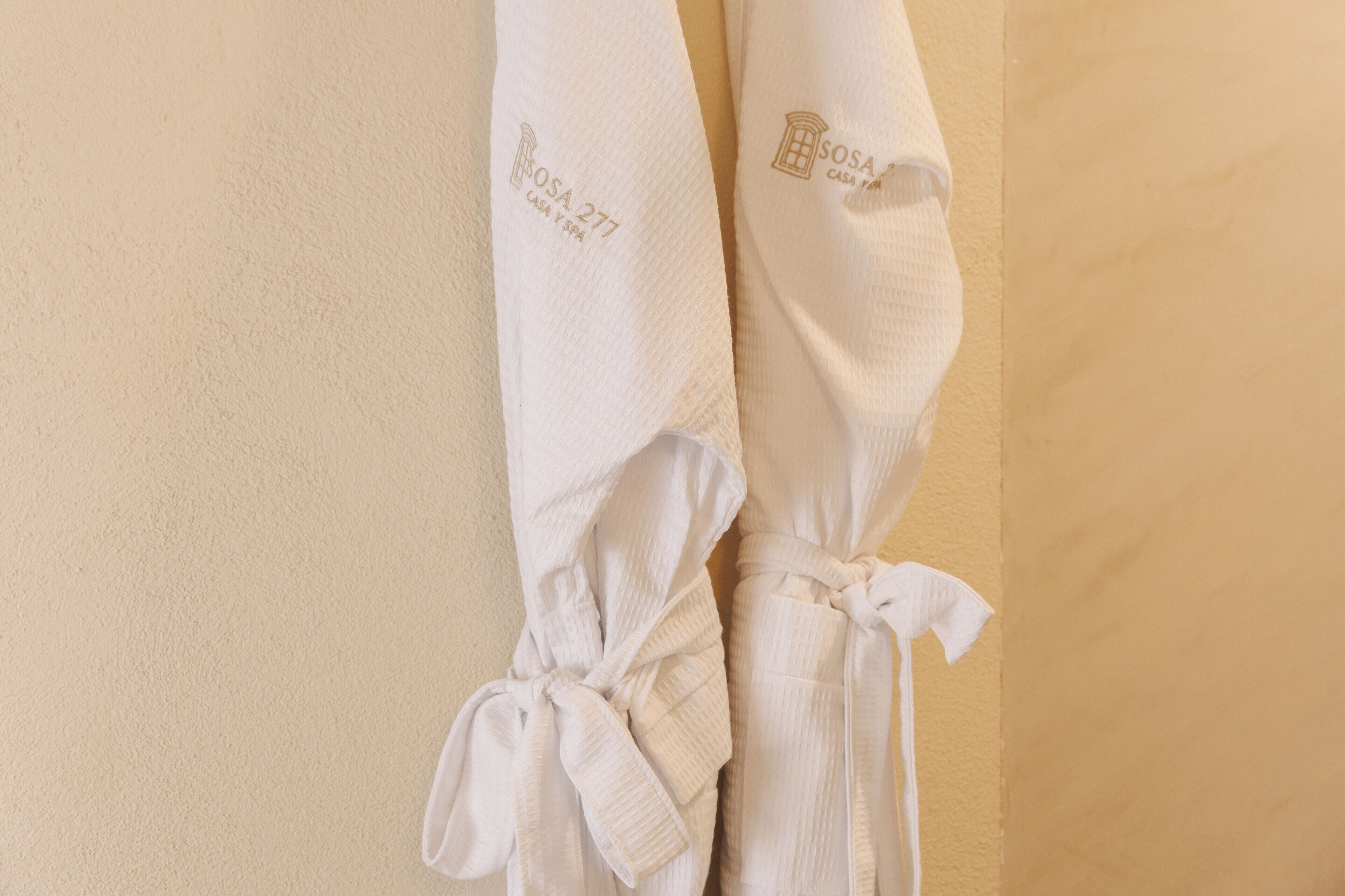 Two white bathrobes hanging on a beige wall, tied with a bow.