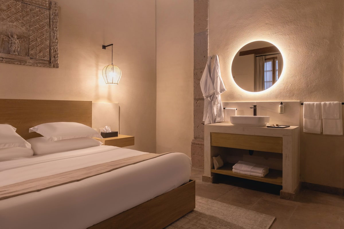Un dormitorio con una cama de madera, una mesita de noche con una lámpara, un espejo redondo sobre el lavabo y un perchero para toallas.